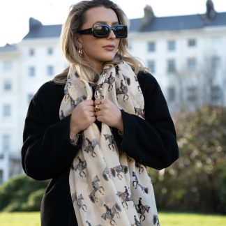 Blonde lady standing outside white buildings in chelsea, wearing an all back outfit, taupe jumper and a hand printed dressage horse design scarf with black sunglasses on