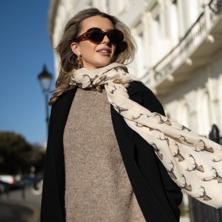 Blonde lady standing outside white buildings in chelsea, wearing an all back outfit, taupe jumper and a hand printed stag heads design scarf with black sunglasses on