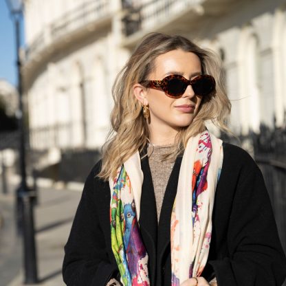 Blonde lady standing outside white buildings in chelsea, wearing an all back outfit, taupe jumper and a hand printed colourful parrots design scarf with black sunglasses on