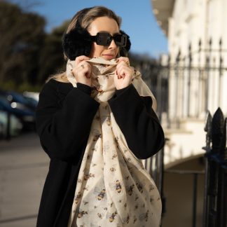 Blonde lady standing outside white buildings in chelsea holding a dark tan leather bag and wearing a hand printed bee design scarf with black sunglasses and earmuffs on