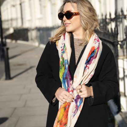 Blonde lady standing outside white buildings in chelsea, wearing an all back outfit, taupe jumper and a hand printed colourful parrots design scarf with black sunglasses on