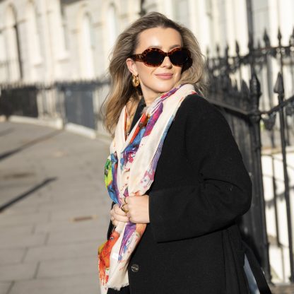 Blonde lady standing outside white buildings in chelsea, wearing an all back outfit, taupe jumper and a hand printed colourful parrots design scarf with black sunglasses on