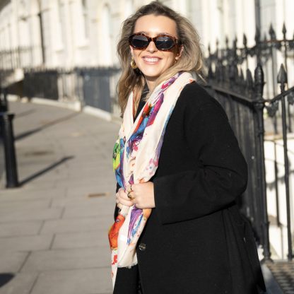 Blonde lady standing outside white buildings in chelsea, wearing an all back outfit, taupe jumper and a hand printed colourful parrots design scarf with black sunglasses on