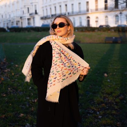 Blonde lady standing outside white buildings in chelsea, wearing an all back outfit, taupe jumper and a hand printed colourful Dachshunds design scarf with black sunglasses on