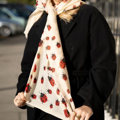 Blonde lady standing outside white buildings in chelsea, wearing an all back outfit, taupe jumper and a hand printed ladybird design scarf with black sunglasses on