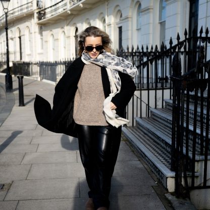 Blonde lady standing outside white buildings in chelsea, wearing an all back outfit, taupe jumper and a hand printed chickens design scarf with black sunglasses on