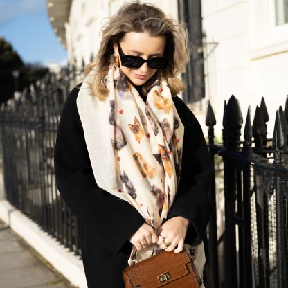 Blonde lady standing outside white buildings in chelsea holding a dark tan leather bag and wearing a hand printed cats and hearts scarf with sunglasses on