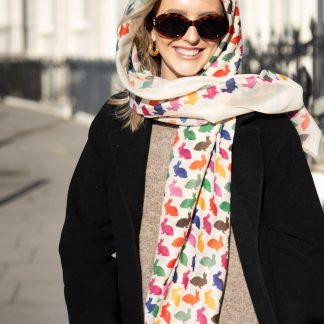 Blonde lady standing outside white buildings in chelsea, wearing an all back outfit, taupe jumper and a hand printed colourful rabbits design scarf with black sunglasses on