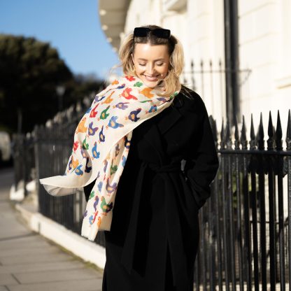 Blonde lady standing outside white buildings in chelsea, wearing an all back outfit, taupe jumper and a hand printed colourful Chickens design scarf with black sunglasses on