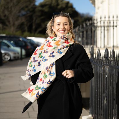 Blonde lady standing outside white buildings in chelsea, wearing an all back outfit, taupe jumper and a hand printed colourful Chickens design scarf with black sunglasses on