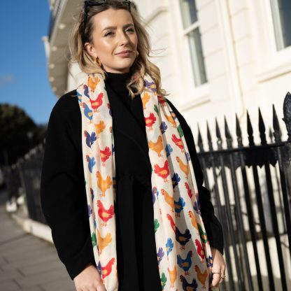 Blonde lady standing outside white buildings in chelsea, wearing an all back outfit, taupe jumper and a hand printed colourful Chickens design scarf with black sunglasses on