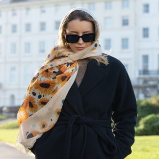 Blonde lady standing outside white buildings in chelsea, wearing an all back outfit, taupe jumper and a hand printed sunflowers design scarf with black sunglasses on