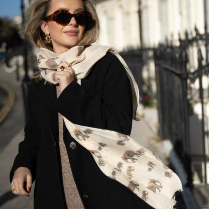 Blonde lady standing outside white buildings in chelsea, wearing an all back outfit, taupe jumper and a hand printed elephants design scarf with black sunglasses on