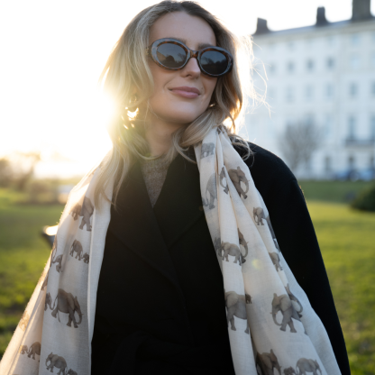 Blonde lady standing outside white buildings in chelsea, wearing an all back outfit, taupe jumper and a hand printed elephants design scarf with black sunglasses on