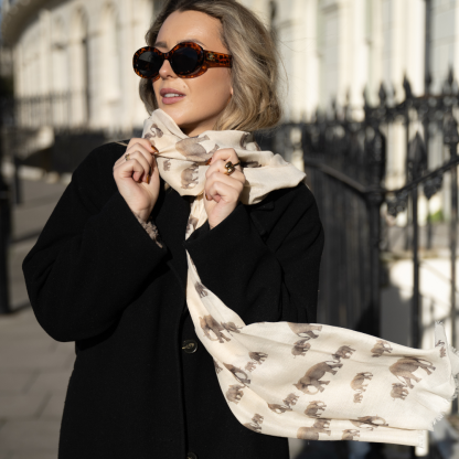 Blonde lady standing outside white buildings in chelsea, wearing an all back outfit, taupe jumper and a hand printed elephants design scarf with black sunglasses on