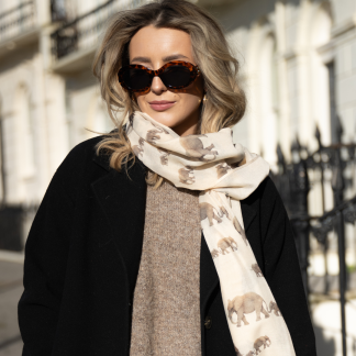 Blonde lady standing outside white buildings in chelsea, wearing an all back outfit, taupe jumper and a hand printed elephants design scarf with black sunglasses on