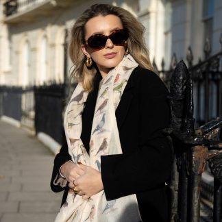 Blonde lady standing outside white buildings in chelsea, wearing an all back outfit, taupe jumper and a hand printed garden birds design scarf with black sunglasses on