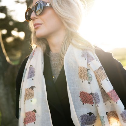 Blonde lady standing outside white buildings in chelsea, wearing an all back outfit, taupe jumper and a hand printed colourful sheep design scarf with black sunglasses on