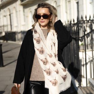 Blonde lady standing outside white buildings in chelsea, wearing an all back outfit, taupe jumper and a hand printed highland Cow design scarf with black sunglasses on