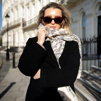 Blonde lady standing outside white buildings in chelsea, wearing an all back outfit, taupe jumper and a hand printed hares design scarf with black sunglasses on