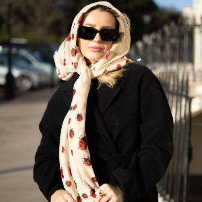 Blonde lady standing outside white buildings in chelsea, wearing an all back outfit, taupe jumper and a hand printed ladybird design scarf with black sunglasses on