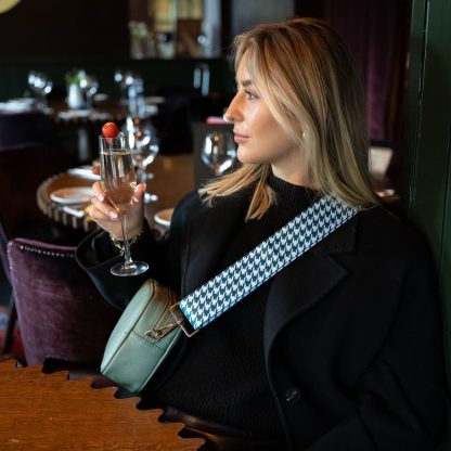 Blonde female model wearing a beautiful crossbody bag strap printed with a teal green dogtooth design sitting in a posh restaurant drinking a glass of champagne