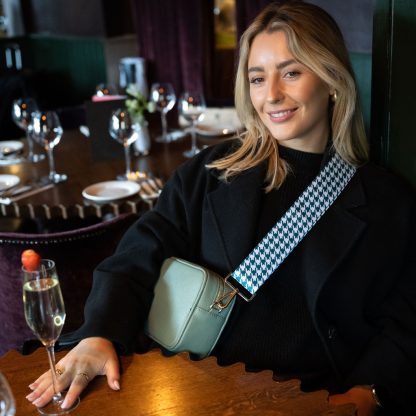 Blonde female model wearing a beautiful crossbody bag strap printed with a teal green dogtooth design sitting in a posh restaurant drinking a glass of champagne