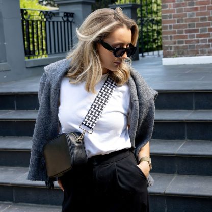 Blonde female model with sunglasses in a minimalist outfit wearing a beautiful crossbody bag strap printed with a black and white dogtooth design standing outside by grey steps