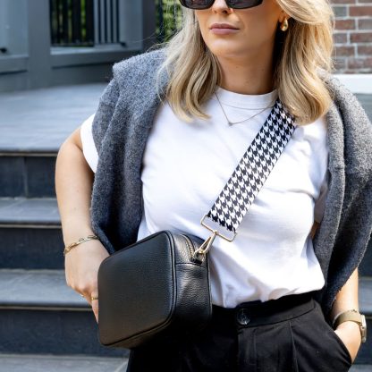 Blonde female model with sunglasses in a minimalist outfit wearing a beautiful crossbody bag strap printed with a black and white dogtooth design standing outside by grey steps