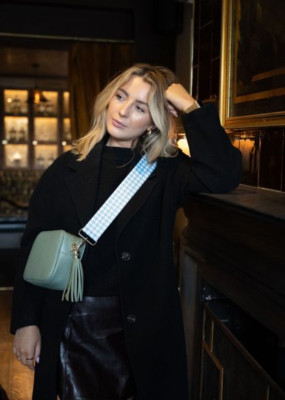 Blonde female model wearing a beautiful crossbody bag strap printed with a mint green dogtooth design sitting in a posh restaurant leaning against a grand fireplace