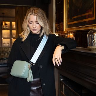 Blonde female model wearing a beautiful crossbody bag strap printed with a mint green dogtooth design sitting in a posh restaurant leaning against a grand fireplace