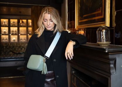 Blonde female model wearing a beautiful crossbody bag strap printed with a mint green dogtooth design sitting in a posh restaurant leaning against a grand fireplace