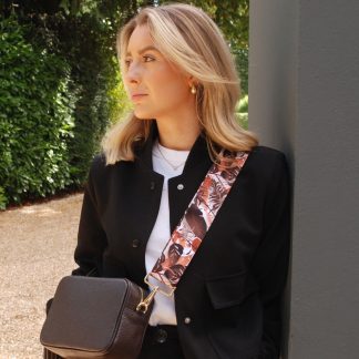 Blonde lady wearing black trousers, white t shirt, grey jumper, black sunglasses with a dark tan italian leather bag and a autumn leaves print bag strap