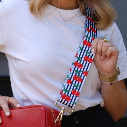 Blonde women wearing cherry bag strap on an italian leather crossbody red bag, white tshirt, gold jewellery and watch