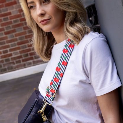 Blonde women wearing cherry bag strap on an italian leather crossbody navy bag, white tshirt, gold jewellery and watch