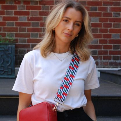 Blonde women wearing cherry bag strap on an italian leather crossbody red bag, white tshirt, gold jewellery and watch