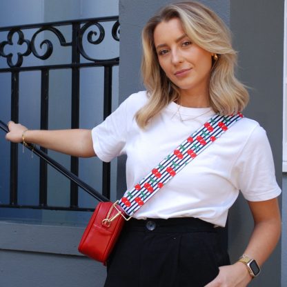 Blonde women wearing cherry bag strap on an italian leather crossbody red bag, white tshirt, gold jewellery and watch