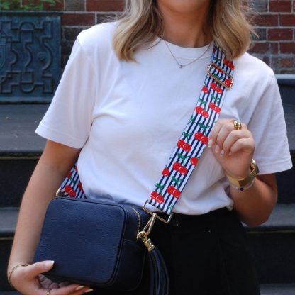 Blonde women wearing cherry bag strap on an italian leather crossbody navy bag, white tshirt, gold jewellery and watch