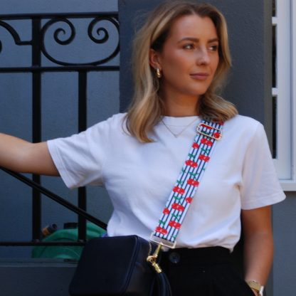 Blonde women wearing cherry bag strap on an italian leather crossbody navy bag, white tshirt, gold jewellery and watch