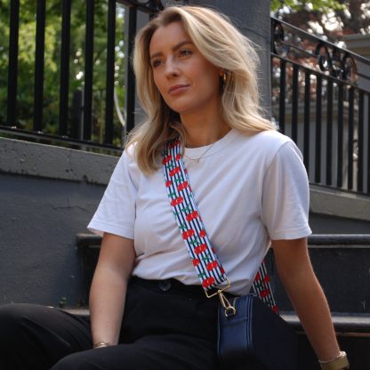 Blonde women wearing cherry bag strap on an italian leather crossbody navy bag, white tshirt, gold jewellery and watch