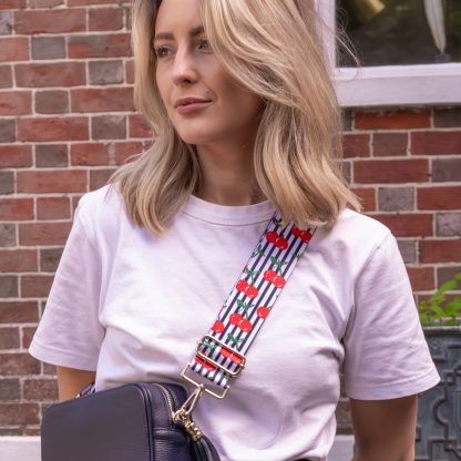 Blonde women wearing cherry bag strap on an italian leather crossbody navy bag, white tshirt, gold jewellery and watch