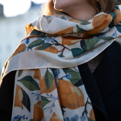 Blonde lady standing outside white buildings in chelsea, wearing an all back outfit, taupe jumper and a hand printed lemons and hydrangeas design scarf with black sunglasses on