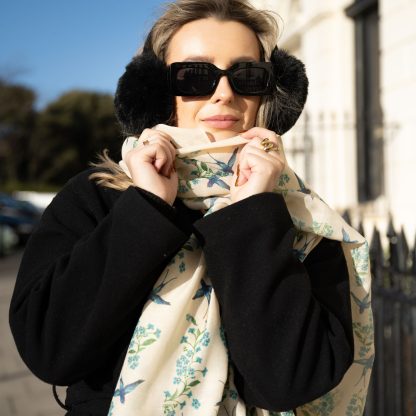 Blonde lady standing outside white buildings in chelsea, wearing an all back outfit, taupe jumper and a hand printed blue birds design scarf with black sunglasses on