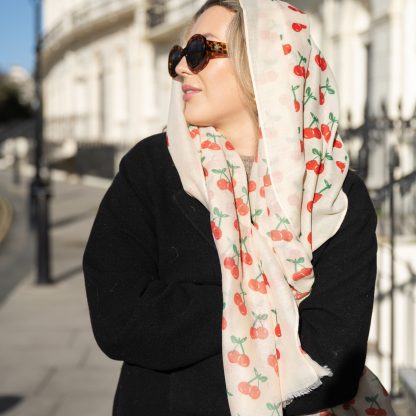 Blonde lady standing outside white buildings in chelsea, wearing an all back outfit, taupe jumper and a hand printed cherries design scarf with black sunglasses on