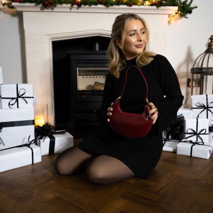 blonde lady sitting in front of fire place with christmas presents holding a burgundy plum red genuine italian leather bag