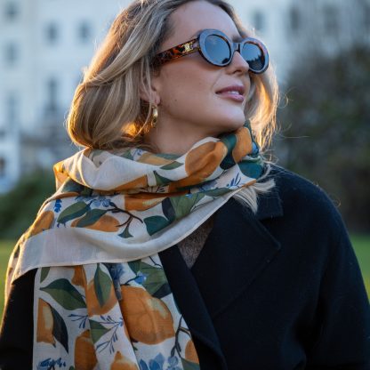 Blonde lady standing outside white buildings in chelsea, wearing an all back outfit, taupe jumper and a hand printed lemons and hydrangeas design scarf with black sunglasses on