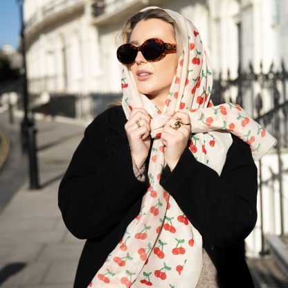 Blonde lady standing outside white buildings in chelsea, wearing an all back outfit, taupe jumper and a hand printed cherries design scarf with black sunglasses on