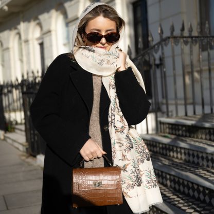 Blonde lady standing outside white buildings in chelsea, wearing an all back outfit, taupe jumper and a hand printed wild ducks design scarf with black sunglasses on