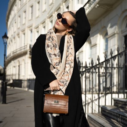 Blonde lady standing outside white buildings in chelsea, wearing an all back outfit, taupe jumper and a hand printed woodland animals design scarf with black sunglasses on