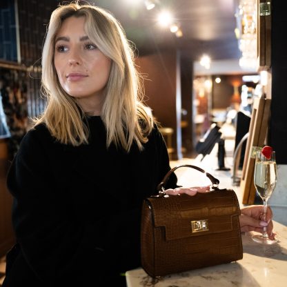 Blonde lady holding tan genuine italian leather croc crossbody bag, standing at a bar in chelsea in black outfit , holding a glass of champagne